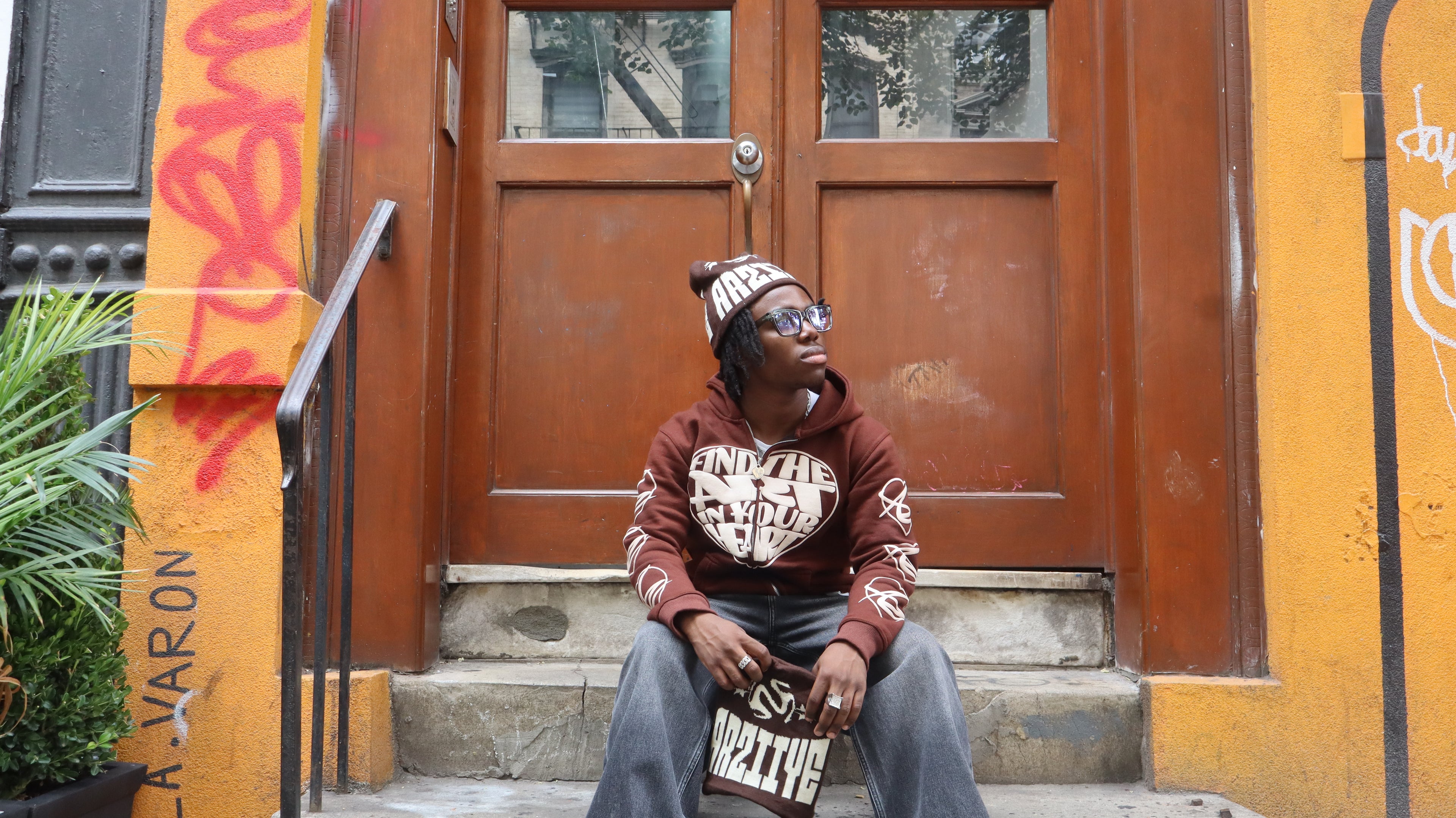 Person sitting on steps in front of a brown door with graffiti, wearing a hoodie and jeans.