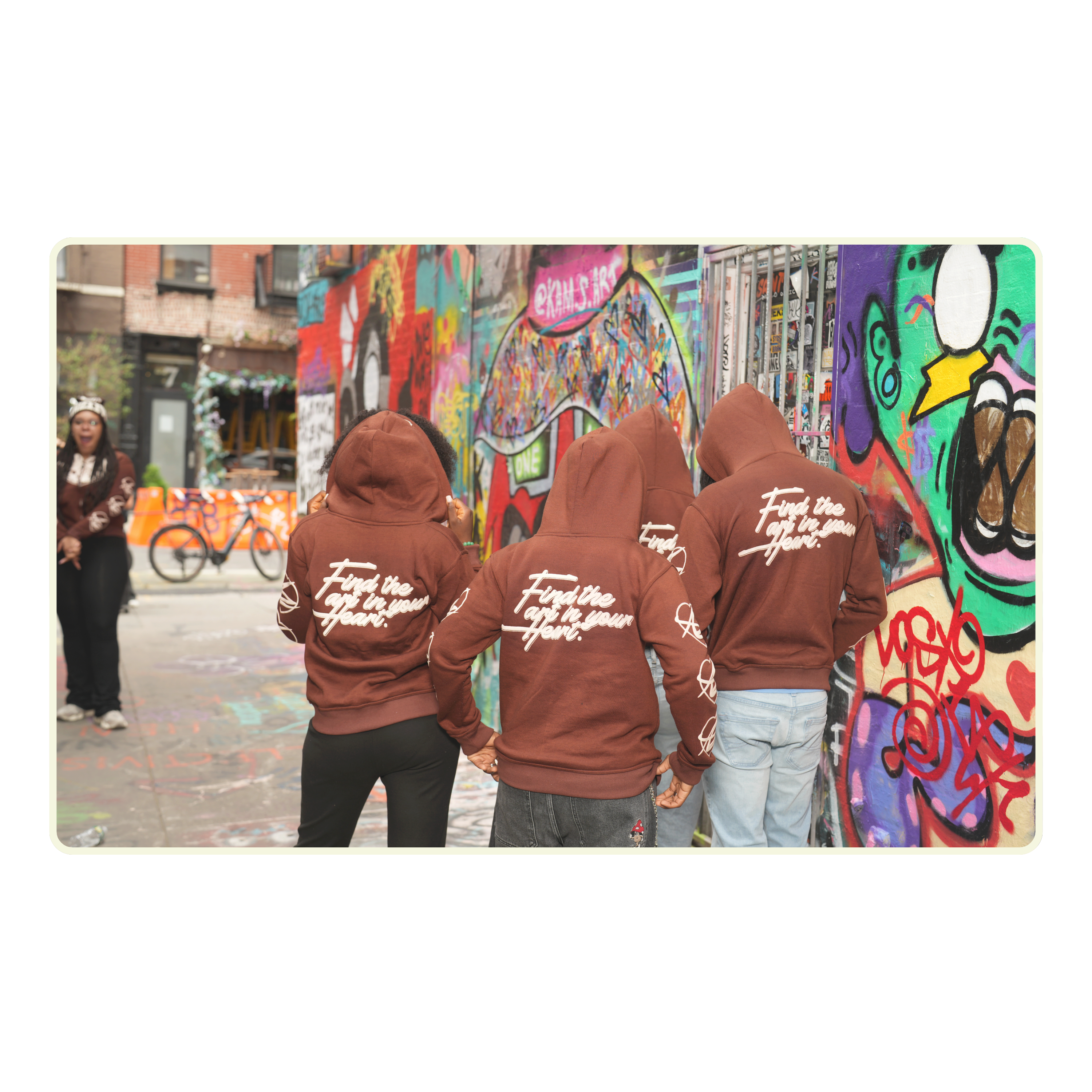 Three people wearing brown hoodies with text, standing in front of a colorful graffiti wall.