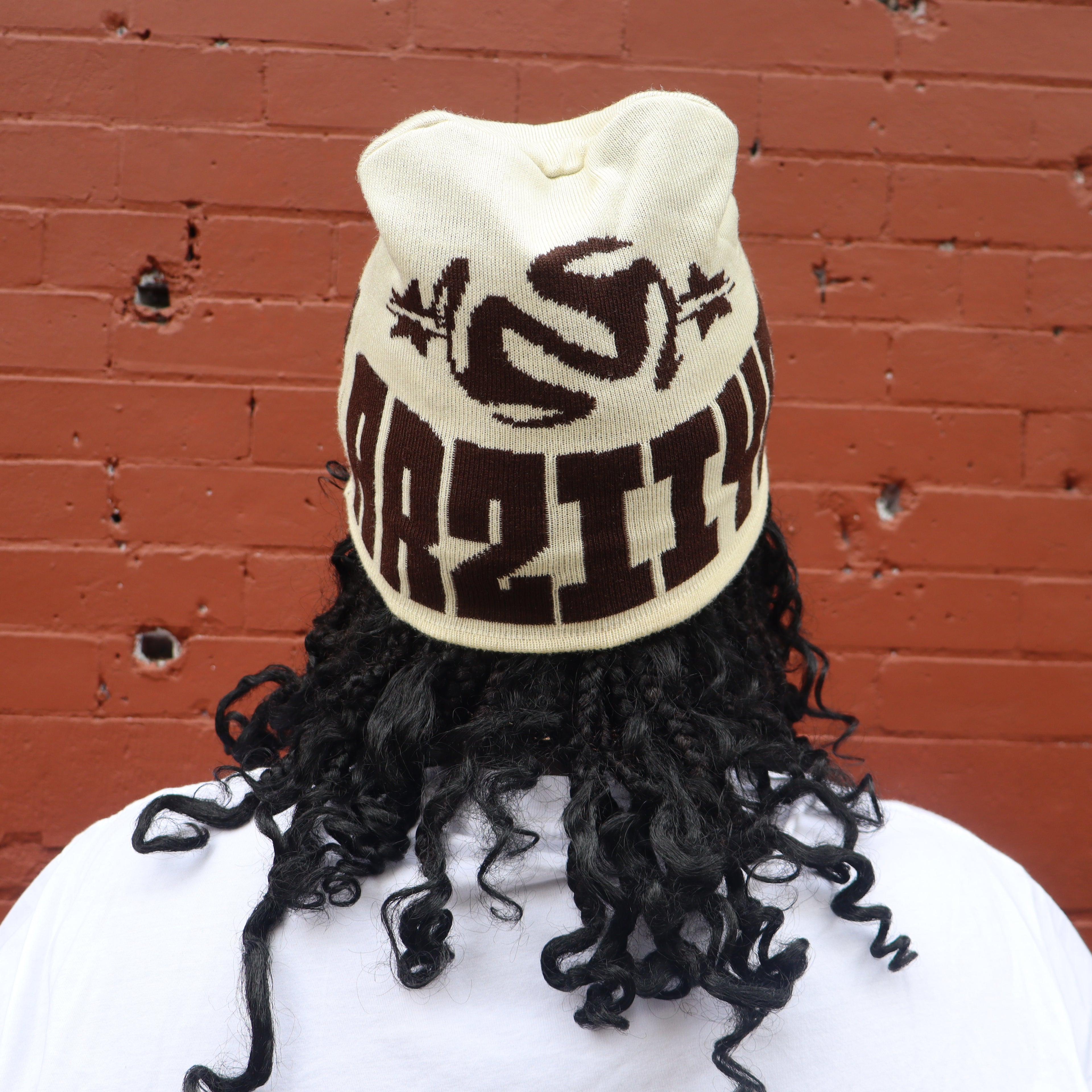 Beanie with black curly hair and brown text against a red brick wall