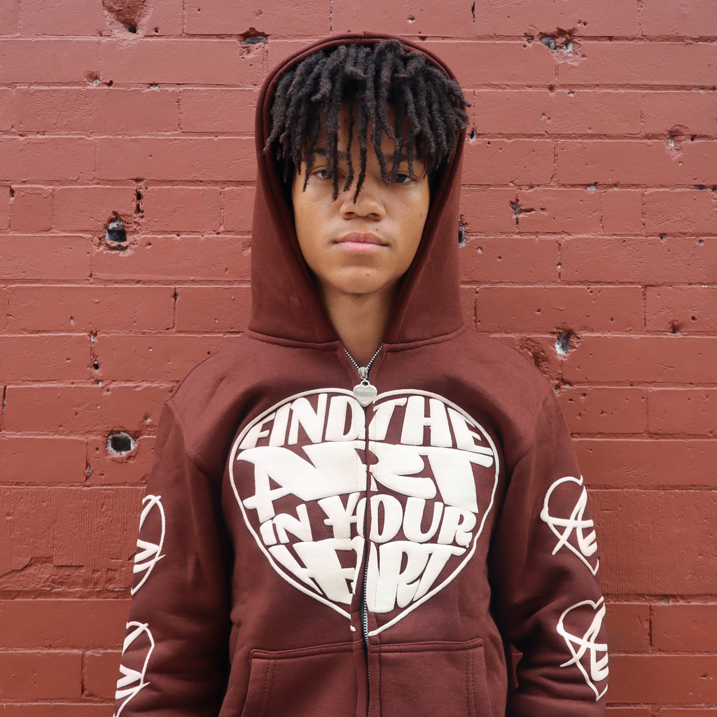 Person wearing a brown hoodie with a heart-shaped text design against a red brick wall.