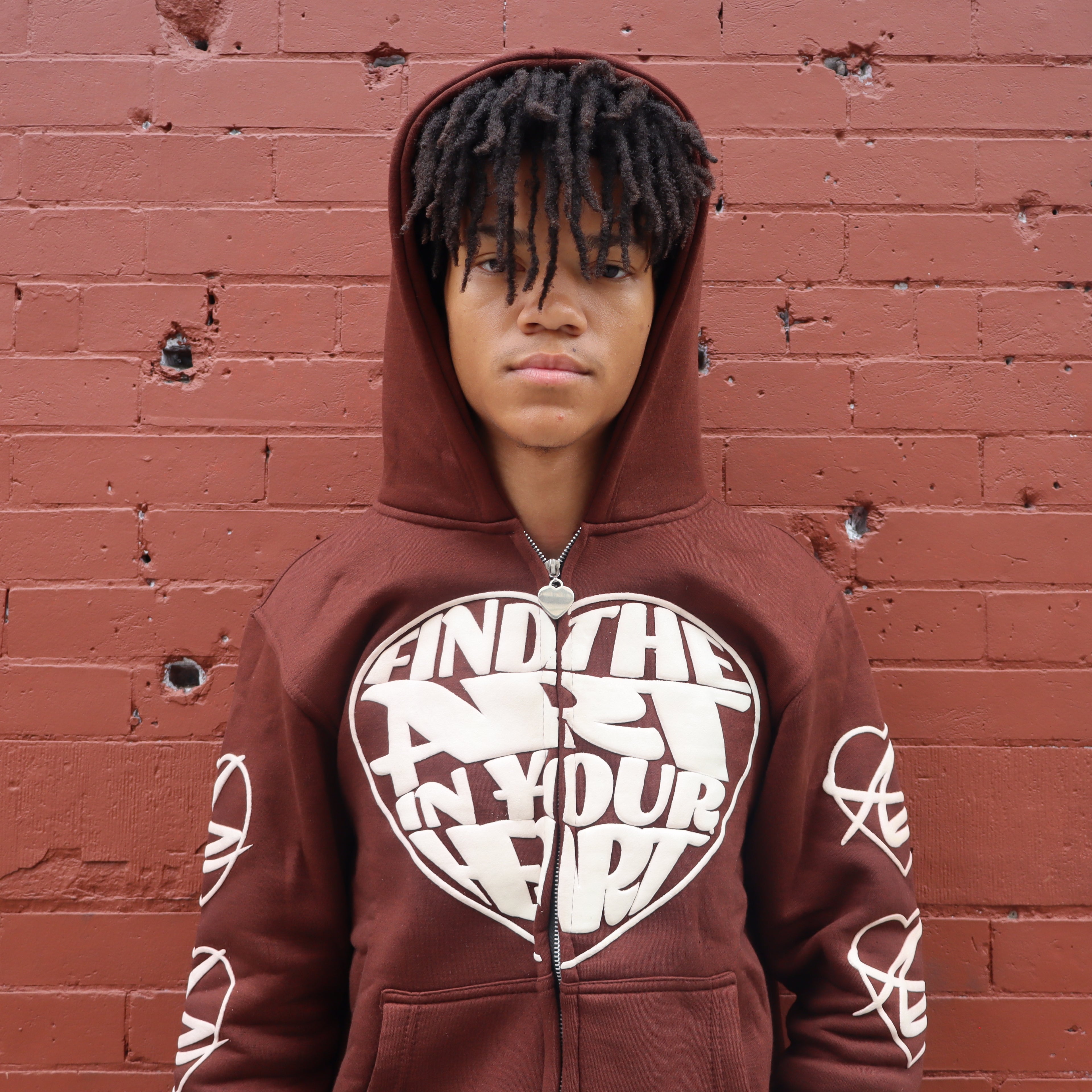 Person wearing a brown hoodie with a heart-shaped text design against a red brick wall.