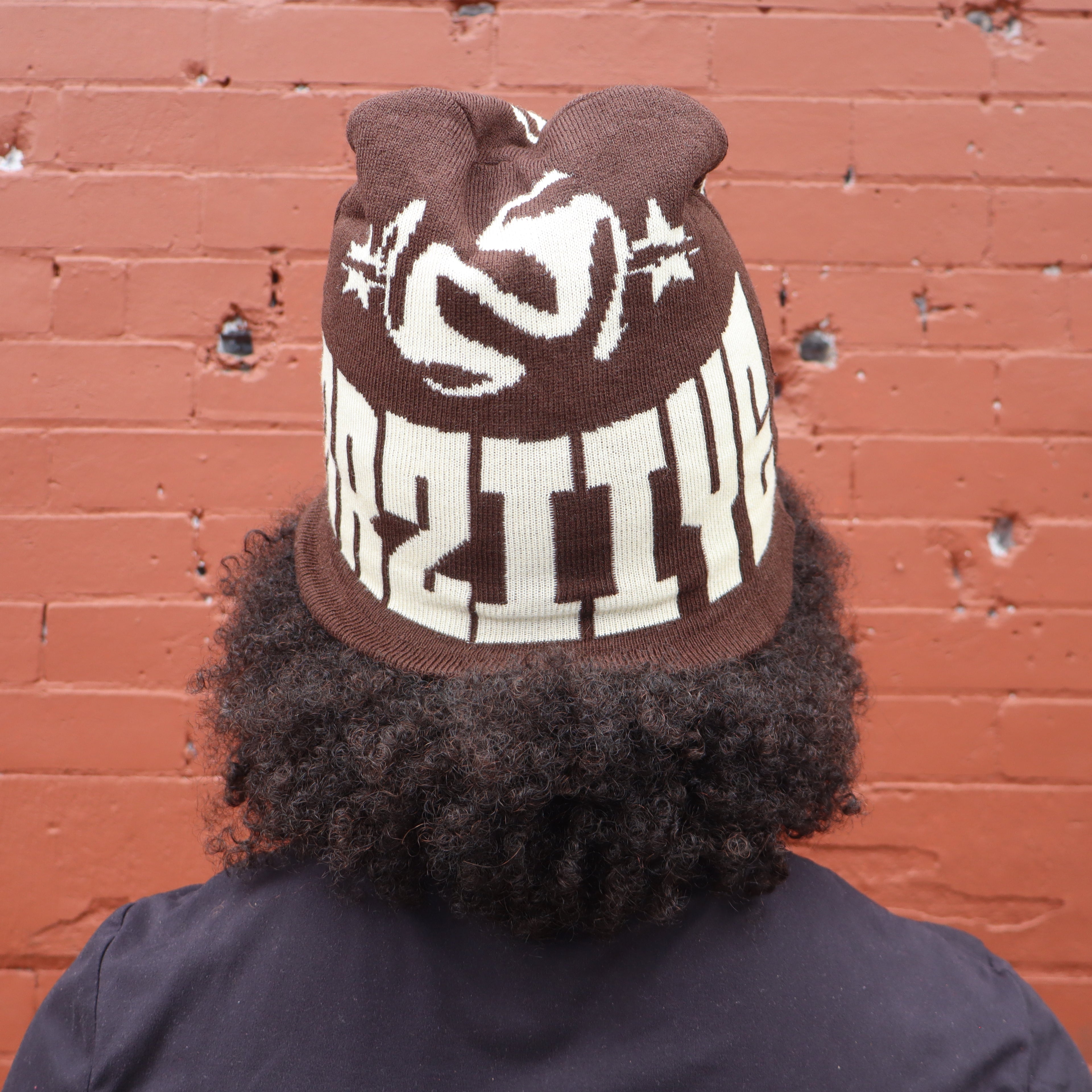 Person wearing a brown knit hat with white text against a red brick wall